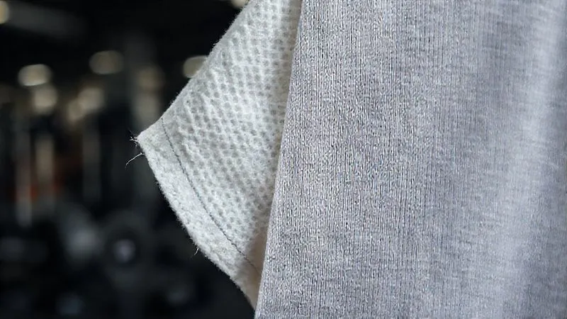 A close-up, photorealistic, professional photography, 8K, cinematic lighting shot of two fabric swatches side-by-side: one a rough, wet cotton, and the other a smooth, dry, high-performance moisture-wicking synthetic blend. Sharp focus on the texture differences, with depth of field blurring a gym background. The image visually contrasts good vs. bad fabric choices for activewear.