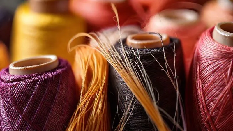 photorealistic, professional photography, 8K, cinematic lighting, sharp focus, depth of field, shot on a high-end DSLR. Close-up of various spools of high-quality silk thread in a gradient of colors, some strands gently pulled and conditioned with a block of beeswax, highlighting the fine texture and readiness for intricate hand-sewing.