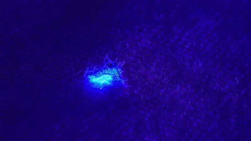 A close-up, macro photorealistic image of a vintage fabric swatch under ultraviolet (UV) light. The original fabric shows a subtle, muted glow, while a small, modern repair patch or re-dye fluoresces brightly in stark contrast. Cinematic lighting emphasizes the UV effect, with sharp focus on the textile fibers. 8K hyper-detailed, shot on a high-end DSLR, illustrating forensic textile analysis.